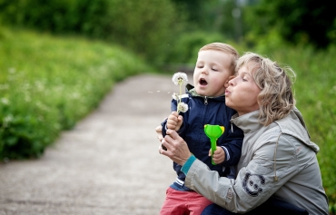 Eine Frau und ein kleiner Junge auf einem Weg in der Natur. Sie pusten gemeinsam einen Pusteblume. Beide haben helle Haut.