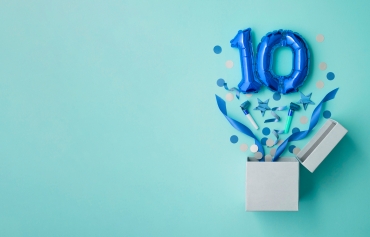 Eine weiße Kiste, aus der blaues Konfetti und blaue Ballons in Form der Zahl 10 auf einem hellblauen Hintergrund
