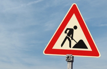 Ein dreieckiges rotes und weißes Verkehrsschild mit einem schwarzen Symbol einer Person, die mit einer Schaufel gräbt. Das Schild ist auf einem Metallpfosten montiert und warnt vor Straßenarbeiten. 