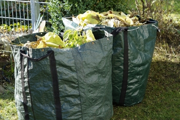 Es sind zwei große, dunkelgrüne Säcke für Gartenabfälle zu sehen. Beide sind mit Blättern, Ästen und anderen Gartenabfällen gefüllt. Sie stehen schräg hintereinander auf grünem Gras, im Hintergrund ist links ein Ausschnitt eines Metallzauns zu erkennen, mittig und rechts ein Teil einer Hecke und Geäst.