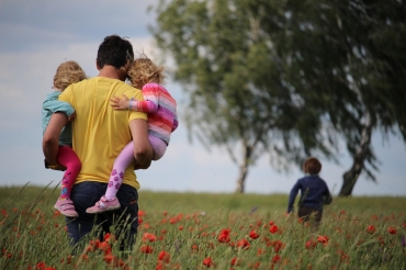 Eine Person in gelbem T-Shirt ist von hinten zu sehen und trägt zwei bunt angezogene Kinder durch eine Wiese mit roten Mohnblumen. Ein anderes Kind rennt voraus.