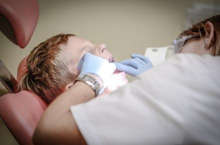 Zahnärztin beugt sich über Jungen, der mit geöffnetem Mund auf dem Behandlungsstuhl liegt. 