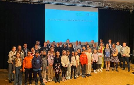 Eine Gruppe verschiedener Menschen, darunter Kinder, stehen auf einer Bühne. Hinter ihnen an der Wand ist auf einer Leinwand eine Präsentation mit dem Titel "Klimaschutzpreis des Landkreises Konstanz 2023" zu sehen.
