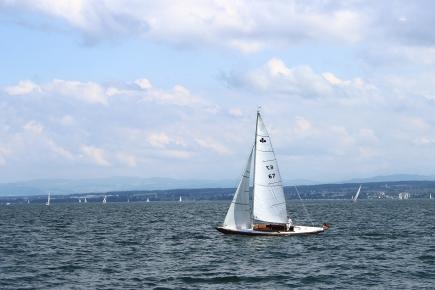 Im Vordergrund ist ein kleines Segelboot auf dem Wasser zu sehen, im Hintergrund ein bewölkter Himmel und kleinere Boote