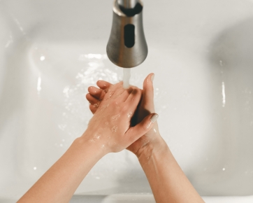 Von oben sind die Hände und Unterarme einer Person mit heller Haut zu sehen. Sie wäscht sich die Hände über einem weißen Waschbecken mit einem silbernen Wasserhahn.