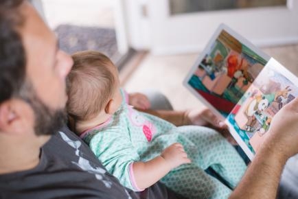 Ein Mann hat ein Baby auf dem Schoß. In einer Hand hält er ein Bilderbuch und zeigt es dem Kind.