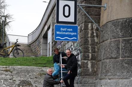 drei Personen stehen an einer Leiter, darüber zwei Schilder