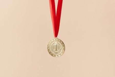 cremefarbener Hintergrund, davor an einem roten Band hängend eine goldene Medaille