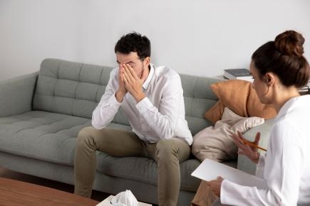 Symbolbild: Mann auf Sofa mit Händen vor dem Gesicht, Frau mit Schreibblock auf Stuhl daneben.