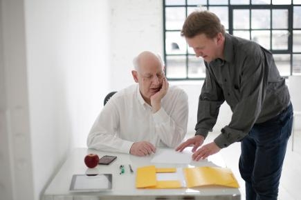 Symbolbild: alter Mann an Tisch mit Unterlagen, nebenstehend erklärt Mann